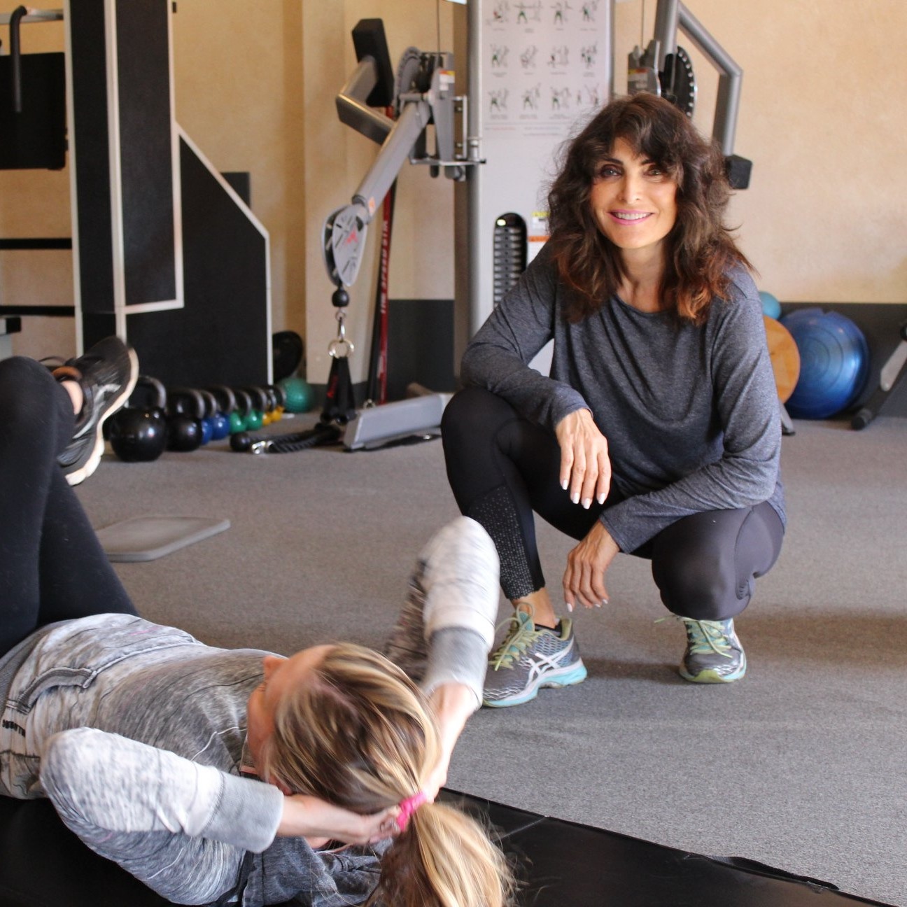 A woman does sit-ups on a gym mat while another woman in athletic wear kneels nearby, smiling, demonstrating strength training for seniors. Gym equipment is visible in the background. A woman does sit-ups on a gym mat while another woman in athletic wear kneels nearby, smiling, demonstrating strength training for seniors. Gym equipment is visible in the background.
