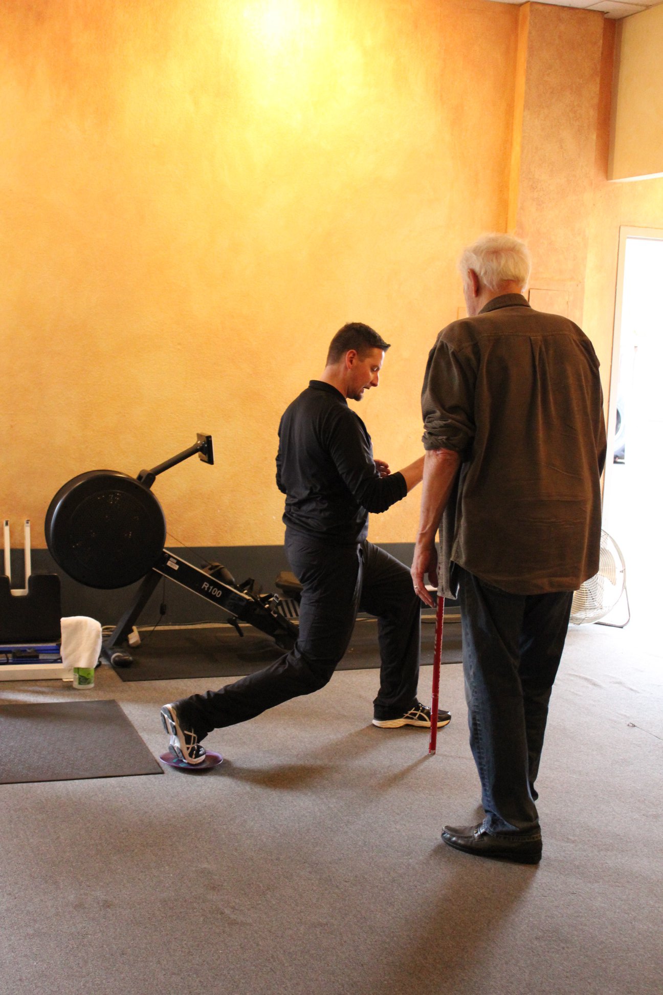 A man performs a lunge exercise with guidance from an older man holding a walking cane, highlighting safe exercises for those with joint issues in a gym setting. A man performs a lunge exercise with guidance from an older man holding a walking cane, highlighting safe exercises for those with joint issues in a gym setting.