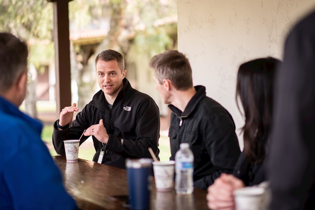 Four people sit at an outdoor wooden table, engaged in conversation about post-surgery recovery. One man gestures while speaking. There are cups, a water bottle, and tumblers on the table.
