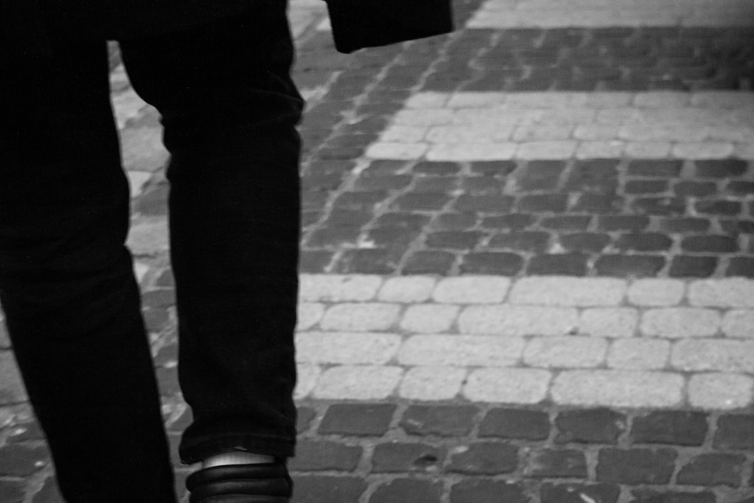 A person showcasing a new approach to fitness walks on a cobblestone pavement, dressed in dark pants and shoes; only their legs and part of a body are visible. The photo is in black and white.