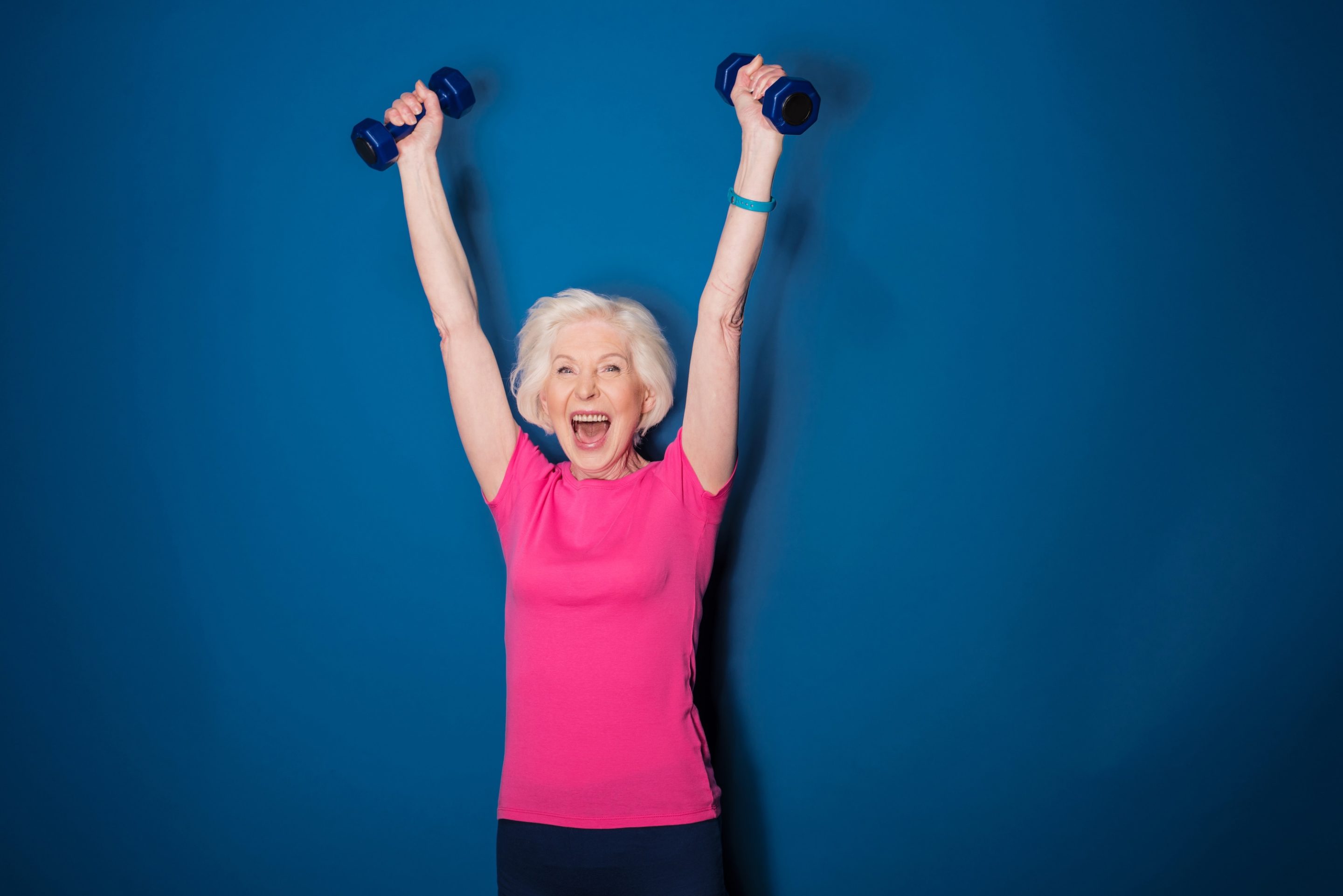 Older woman in a pink shirt smiles and raises both arms overhead, each holding a blue dumbbell, standing against a solid blue background, embodying aging gracefully and the benefits of muscle memory.