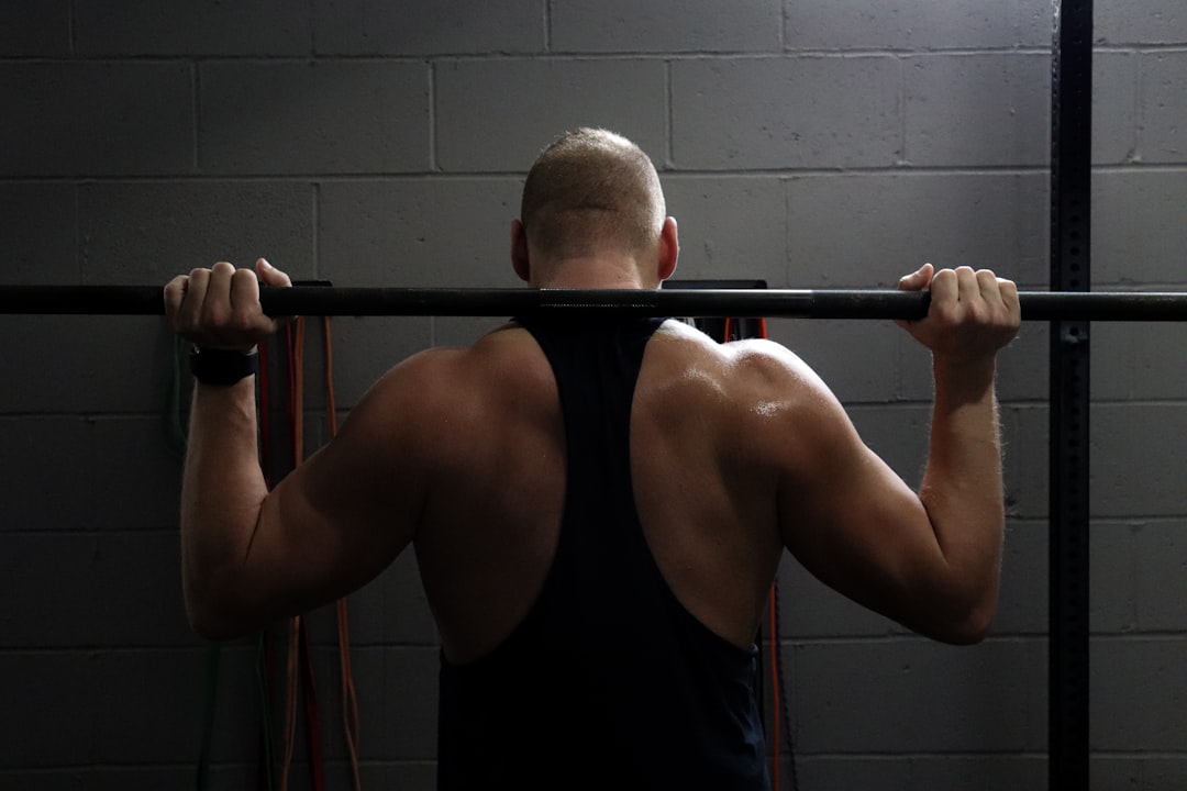 A person in a tank top faces a gray cinder block wall while holding a barbell across their shoulders in a gym setting, focused on strength training and ignoring muscle loss and associated health risks.