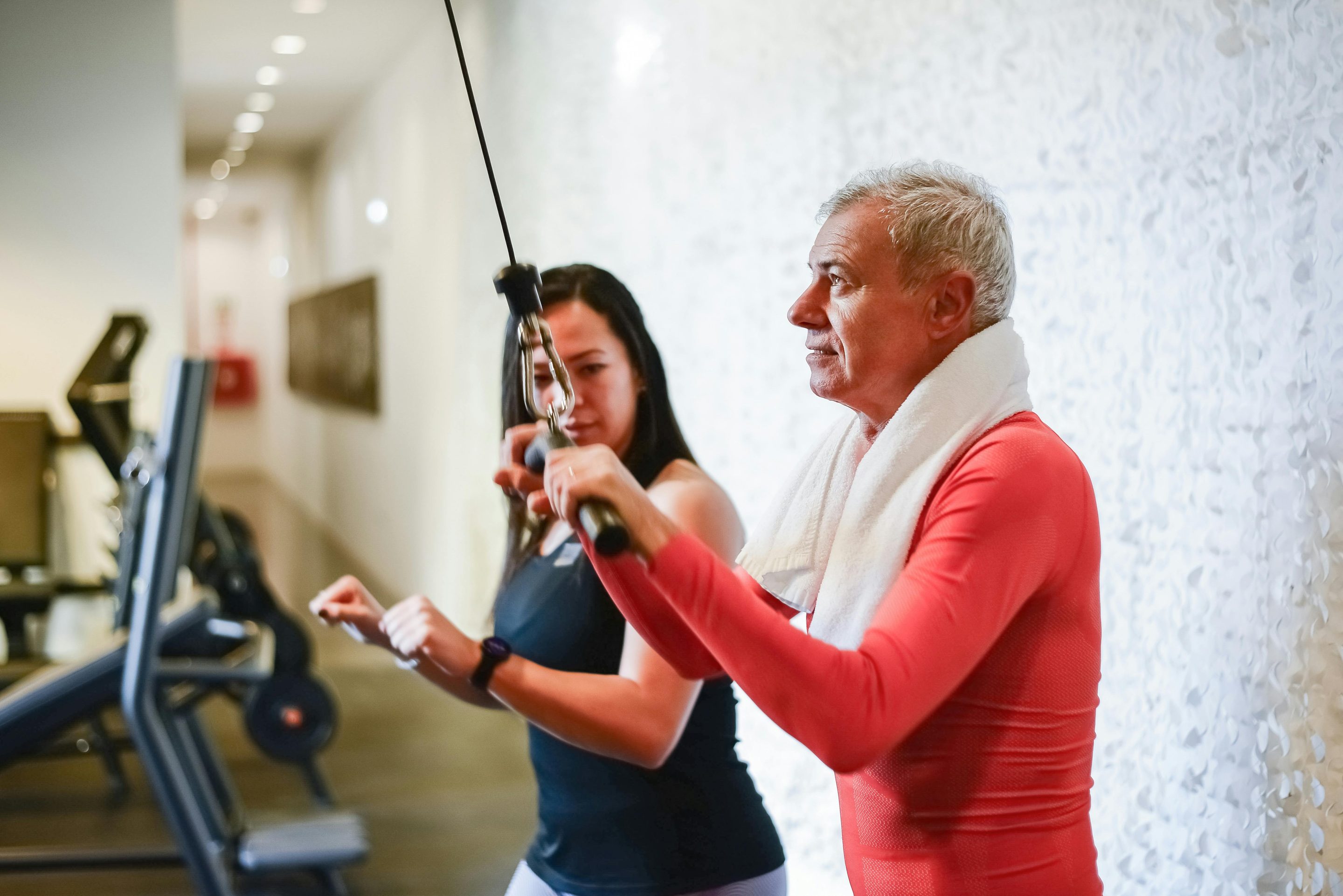 An older man works to prevent muscle loss on a cable machine with a towel over his shoulders, while a woman in sportswear coaches him at the gym.