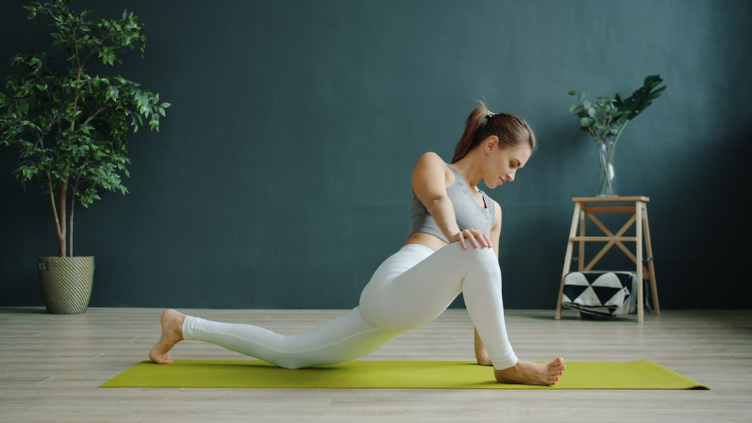 Woman doing a low lunge stretch on a green yoga mat in a room with wooden floor, a potted plant, and a small table with decor—perfect exercises to support joint health.