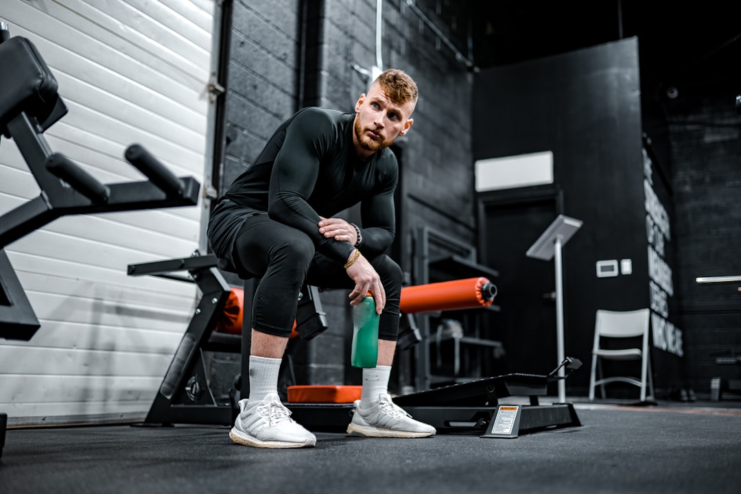 Man in athletic wear sits on gym equipment holding a green water bottle, surrounded by fitness machines in a modern gym setting, focused on building muscle and promoting healthy aging.