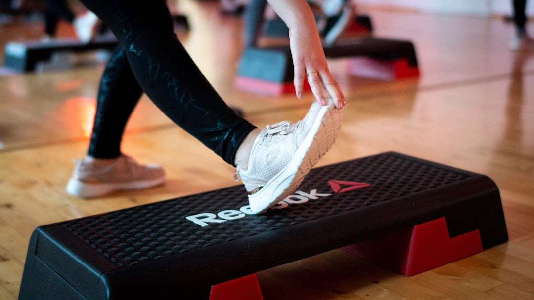 Person restarting fitness after 50 stretches by placing one foot on a Reebok aerobic step platform in a gym, with others exercising in the background.