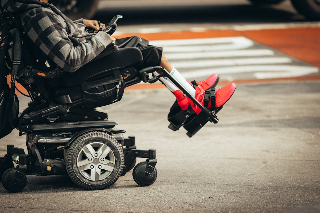 A person in a motorized wheelchair, showcasing independence and mobility, waits at a crosswalk, wearing red shoes and a striped hoodie.