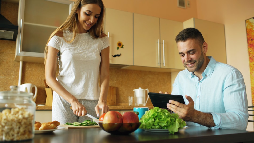 A woman slices vegetables in a kitchen while a man sits at the table using a tablet, perhaps researching DIY diet changes. Fresh produce and a jar of cereal are on the table, creating an inviting scene focused on healthy nutrition. A woman slices vegetables in a kitchen while a man sits at the table using a tablet, perhaps researching DIY diet changes. Fresh produce and a jar of cereal are on the table, creating an inviting scene focused on healthy nutrition.